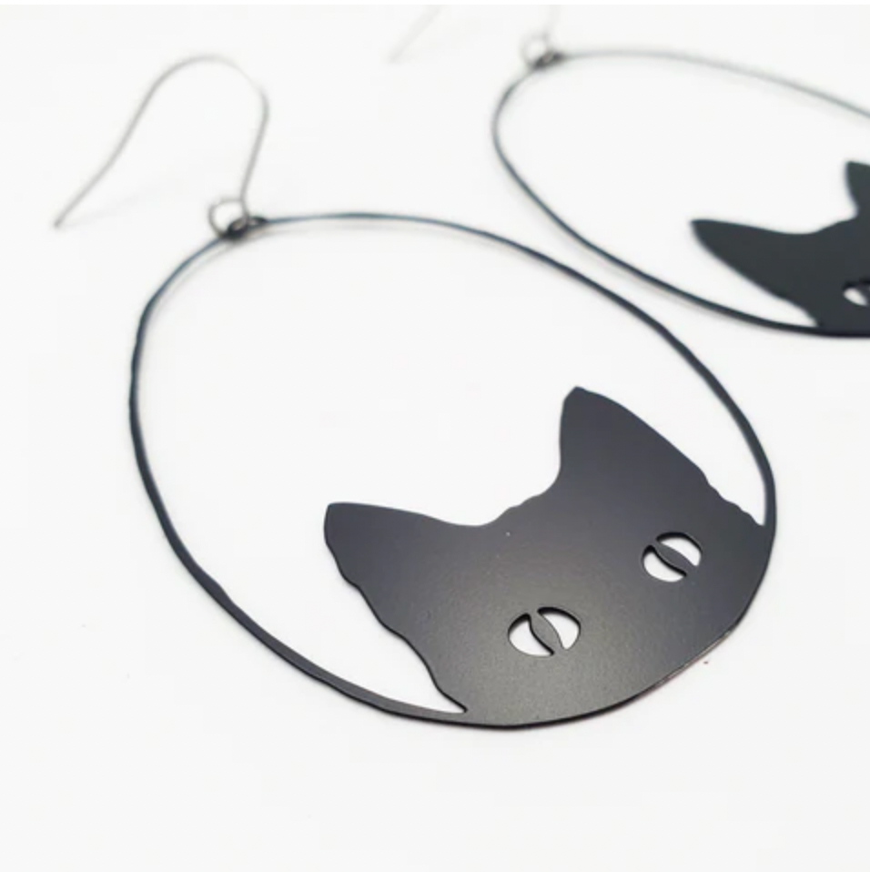 Close-up of the Denz Cat Black Earrings by Blue Dog Posters, a pair of lightweight statement earrings shaped like cat's heads and crafted from recycled stainless steel. The design features a simple silhouette of cat ears and eyes within an oval frame. These unique pieces, handmade in Noosa, are set against a plain white background.