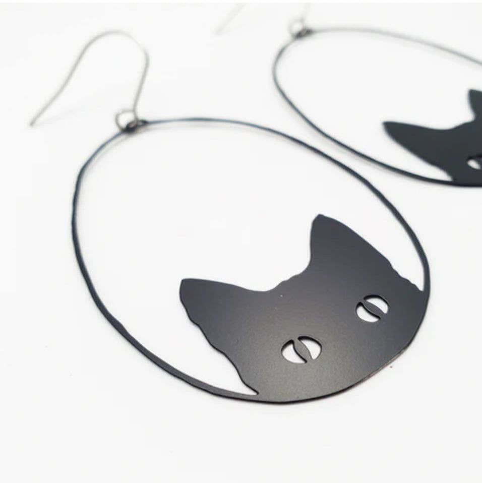 Close-up of the Denz Cat Black Earrings by Blue Dog Posters, a pair of lightweight statement earrings shaped like cat's heads and crafted from recycled stainless steel. The design features a simple silhouette of cat ears and eyes within an oval frame. These unique pieces, handmade in Noosa, are set against a plain white background.