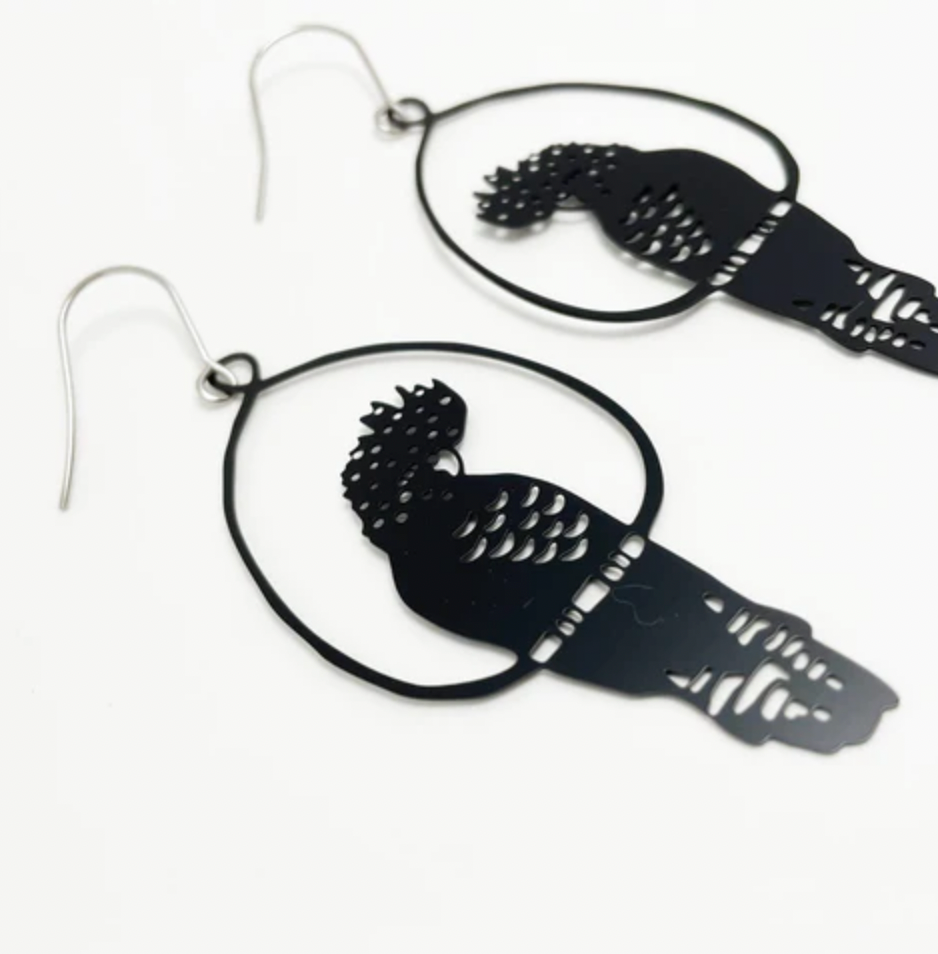 A close-up view of two Black Denz Cockatoo Earrings by Blue Dog Posters, each dangling within an open circular frame. Handmade in Noosa from recycled stainless steel, these super lightweight statement earrings feature intricate cut-out feather details and are attached to metal hooks. The white background makes the earrings the focal point.