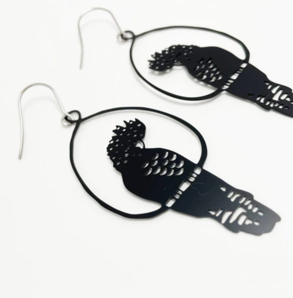 A close-up view of two Black Denz Cockatoo Earrings by Blue Dog Posters, each dangling within an open circular frame. Handmade in Noosa from recycled stainless steel, these super lightweight statement earrings feature intricate cut-out feather details and are attached to metal hooks. The white background makes the earrings the focal point.