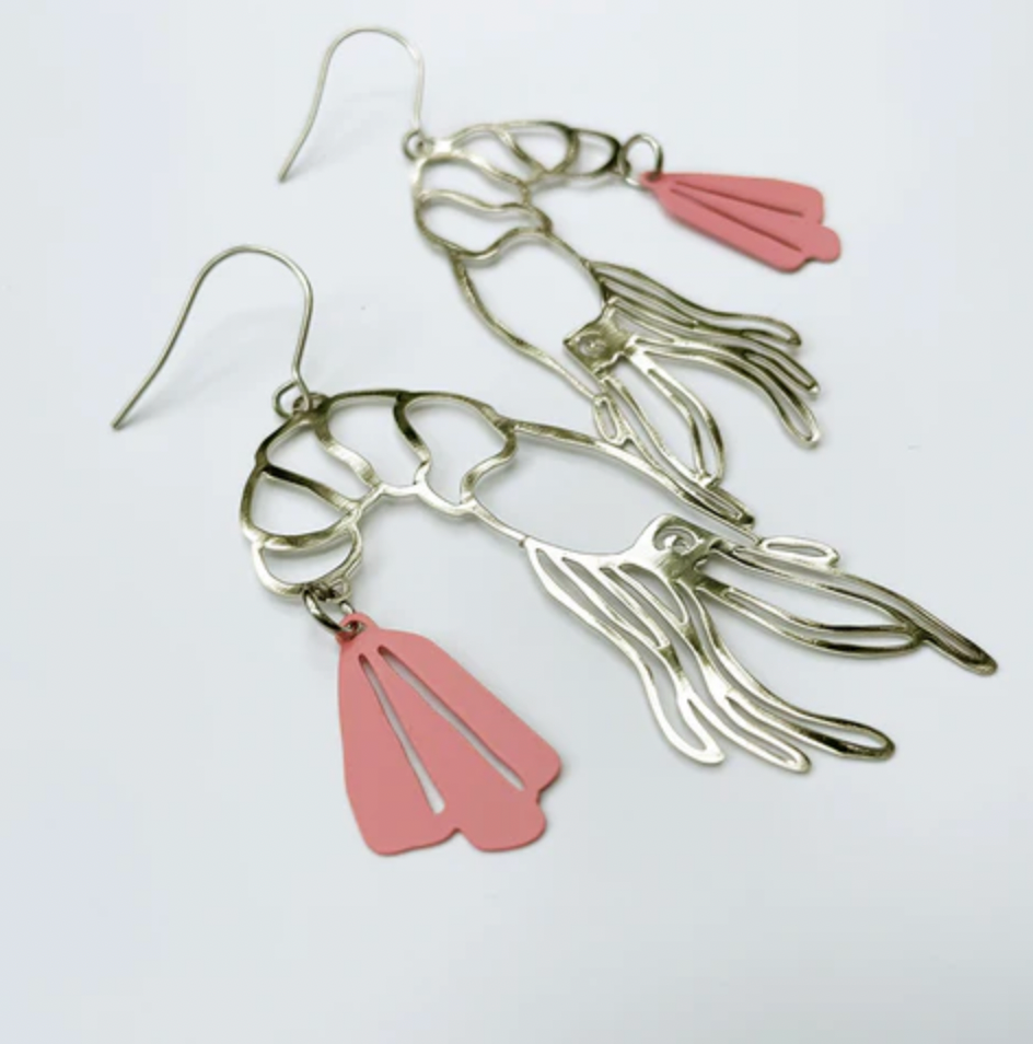 The Denz Prawn Dangles Pink Earrings from Blue Dog Posters are a unique, lightweight pair of statement earrings, intricately crafted to resemble squid. They feature detailed metalwork in a silver hue with striking pink accents on the tail fins and are photographed against a white background. These earrings are handmade in Noosa from recycled stainless steel and come with hook-style backs for easy wearing.