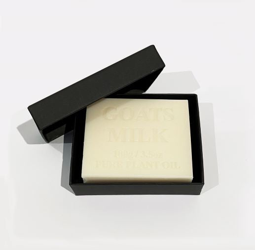 A white soap bar labeled "Hustle and Co Soap - Mansplaining" by Blue Dog Posters is partially visible in an open black box against a plain white background, showcasing natural skincare at its finest.