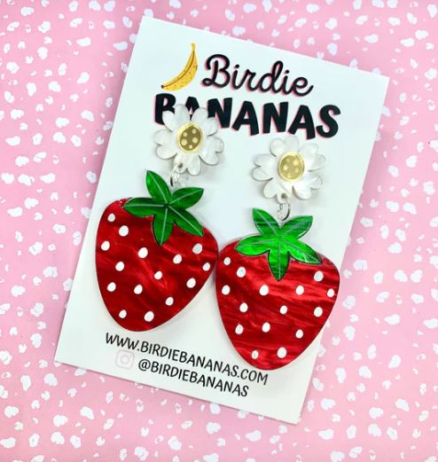 A pair of "Strawberry Red Acrylic Earrings Blue" by Blue Dog Posters. These fun, quirky accessories feature white flowers with yellow centers at the top and large red strawberries with white polka dots hanging below. The background is a pink surface with white dots, perfectly showcasing this delightful piece of hand-made jewelry.