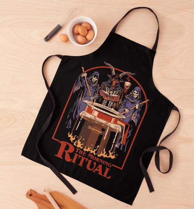 A black Steven Rhodes Morning Ritual Apron from Blue Dog Posters lies flat on a wooden surface, part of our Homewares Collection. The apron features a colorful illustration of three skeleton figures in robes performing a ritual around a bubbling cauldron, with the text "The Morning Ritual" below. Nearby are eggs in a bowl and wooden utensils. It's machine washable for easy care.