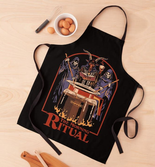 A black Steven Rhodes Morning Ritual Apron from Blue Dog Posters lies flat on a wooden surface, part of our Homewares Collection. The apron features a colorful illustration of three skeleton figures in robes performing a ritual around a bubbling cauldron, with the text "The Morning Ritual" below. Nearby are eggs in a bowl and wooden utensils. It's machine washable for easy care.
