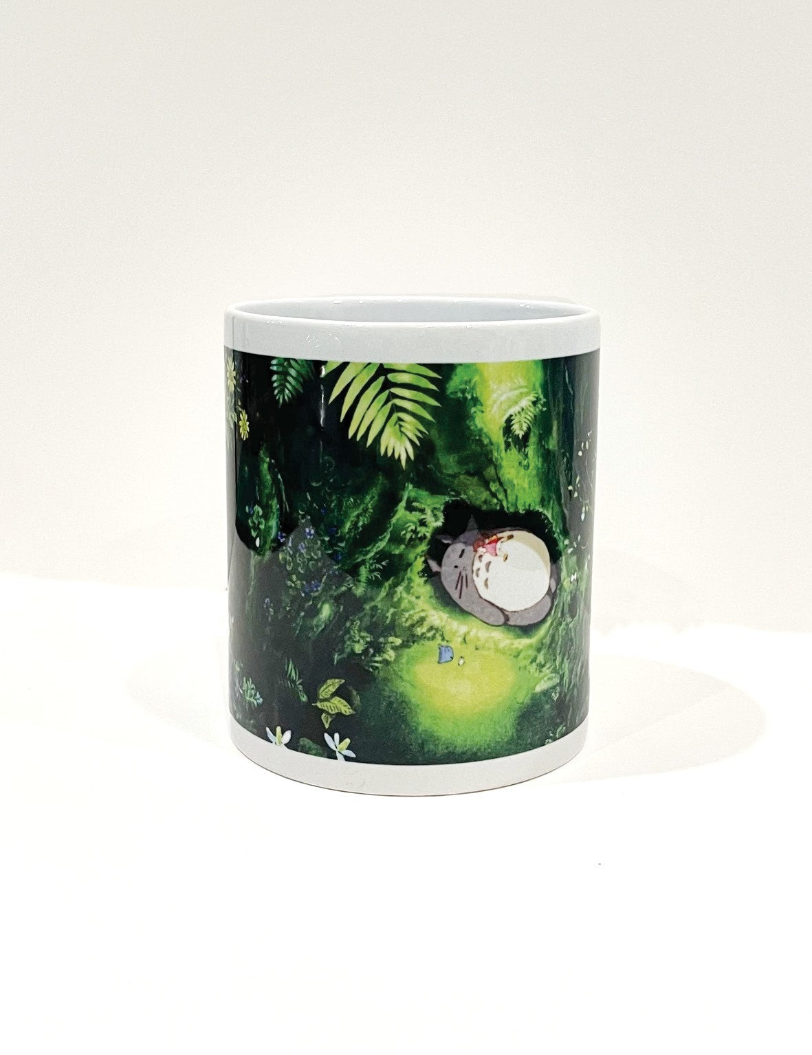 Part of our Homewares Collection, this white mug, "Ghibli - Totoro Nest" by Blue Dog Posters, features a detailed illustration of a small sleeping cat nestled in a lush green forest with ferns, moss, and foliage. The background blends soft greens, creating a serene atmosphere. Plus, it's dishwasher safe for easy cleaning.