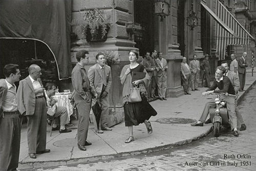 A woman in a knee-length dress confidently walks down a sidewalk in Italy, 1951, while several men standing and sitting nearby observe her. This candid street scene in black and white is captured on premium 150gsm gloss paper for an American Girl In Italy 1951 by IMP 091. Credit: Ruth Orkin, "American Girl in Italy 1951.