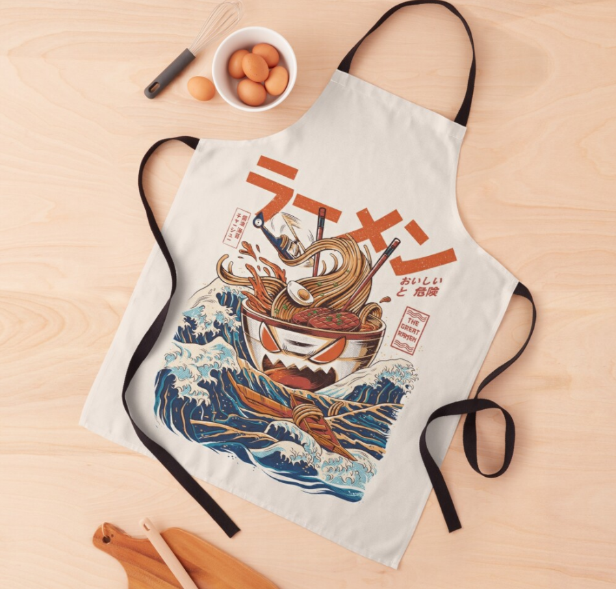 This Blue Dog Posters The Great Ramen Apron Cream features a colorful illustration of a bowl of ramen with chopsticks. The design includes waves and various Japanese text elements. It is machine washable for easy care and laid out flat on a light wooden surface, alongside a whisk with eggs and a wooden spoon.