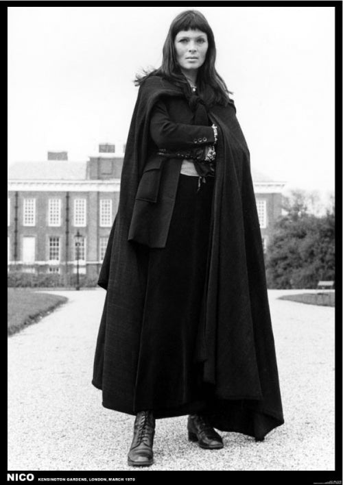 A person stands outdoors wearing a long dark cloak in front of a historic building. They have long hair and a serious expression, with arms crossed. The black-and-white image, reminiscent of a classic rock poster, was taken in Kensington Gardens, London, March 1970. This captures the essence of The Velvet Underground & Nico by MUS 054.