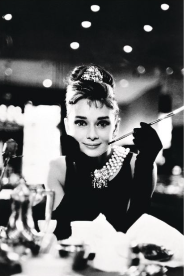 Black and white photo of a woman with an updo hairstyle and a pearl necklace, wearing gloves and holding a long cigarette holder. She is sitting at a table with dinnerware, looking towards the camera with a slight smile. This Audrey Hepburn by IMP 088, printed on premium 150gsm gloss paper, features blurred lights in the background.