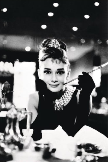 Black and white photo of a woman with an updo hairstyle and a pearl necklace, wearing gloves and holding a long cigarette holder. She is sitting at a table with dinnerware, looking towards the camera with a slight smile. This Audrey Hepburn by IMP 088, printed on premium 150gsm gloss paper, features blurred lights in the background.