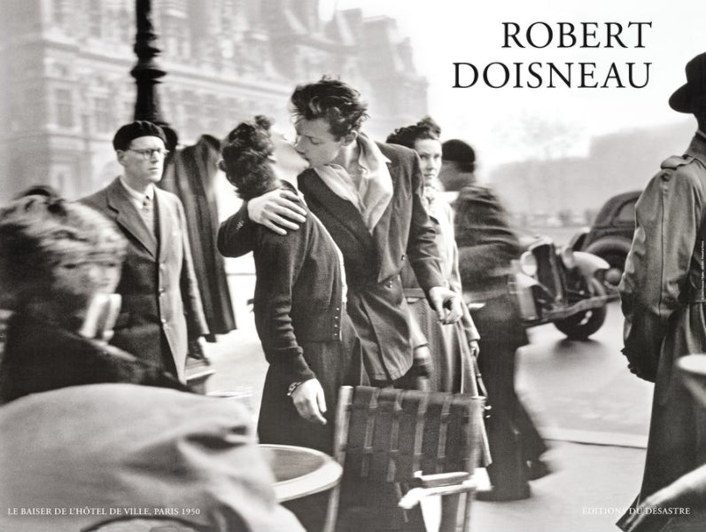 A black and white photo by Robert Doisneau shows a man and woman kissing passionately on a busy Paris street in 1950. Bystanders walk past, some glancing at the couple. This iconic scene captures a mix of motion and stillness in a bustling city atmosphere, perfect for an IMP 087 Le Baiser de L Hotel de Ville Art Print.