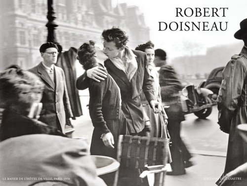 A black and white photo by Robert Doisneau shows a man and woman kissing passionately on a busy Paris street in 1950. Bystanders walk past, some glancing at the couple. This iconic scene captures a mix of motion and stillness in a bustling city atmosphere, perfect for an IMP 087 Le Baiser de L Hotel de Ville Art Print.
