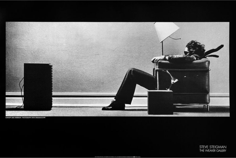 A person sits in an armchair tilted back by a powerful gust of air from a speaker in front of them, hair and clothing blown back dramatically. A stylish floor lamp and side table accentuate the scene, captured on premium 150gsm gloss paper. The image, printed extra large, is in black and white. This is the BLOWN AWAY POSTER ART PRINT from IMP 089.
