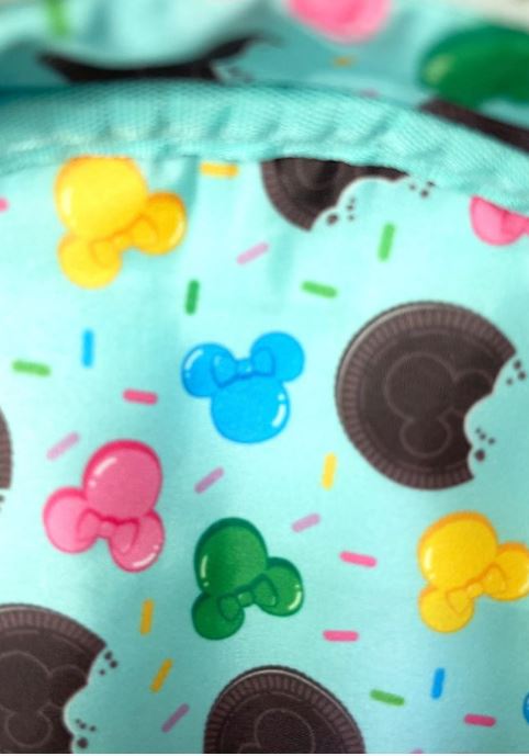 A close-up view of the Disney "Minnie Ice Cream Backpack" by Blue Dog Posters reveals a vibrant fabric pattern with blue, yellow, pink, and green Mickey Mouse silhouettes, chocolate cookies, and multicolored sprinkles against a light turquoise background. Perfect for any Loungefly Backpack collector or Disney aficionado.