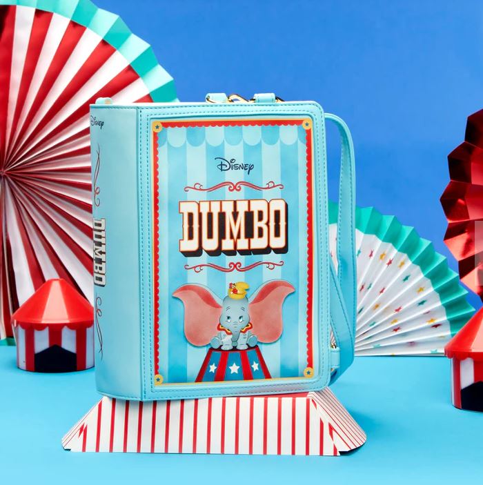 The Dumbo 1941 Book Convertible Crossbody by Blue Dog Posters features Disney's Dumbo on the front, showcasing his charming face and ears. The background is a striking blue adorned with red and white fan decorations and circus-themed elements, including a tiny red and white circus tent at the bottom.