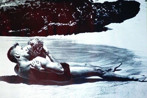 Black and white image of two people lying on a sandy beach. They are passionately kissing, with the person on the left leaning over the person on the right. Dark rocks can be seen in the background. This scene is beautifully captured on premium gloss paper, ideal for From Here to Eternity aficionados from IMP.