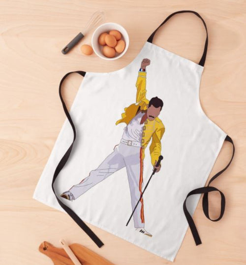 A white apron from our homewares collection, the Freddie Mercury Apron by Blue Dog Posters, features a graphic of a person in a yellow jacket and white pants striking a dynamic pose with a microphone stand. Laid on a light wooden surface beside kitchen essentials, this machine washable apron is perfect for adding flair to your cooking routine.