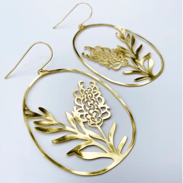A pair of Denz Grevilleas Gold Earrings from Blue Dog Posters featuring an oval-shaped design with intricate floral cutouts. These lightweight statement earrings showcase a detailed flower with leaves in the center of the oval, hanging from simple gold hooks. The background is white.