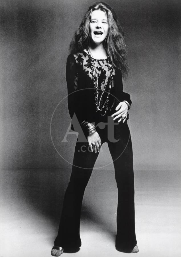 Black and white photo of Janis Joplin with long, wavy hair wearing a dark outfit with intricate designs on the top. She stands against a plain background, laughing expressively with her mouth open wide and her right hand resting near her thigh. Perfect for a rock poster printed on premium 150gsm gloss paper from the MUS 294 collection.