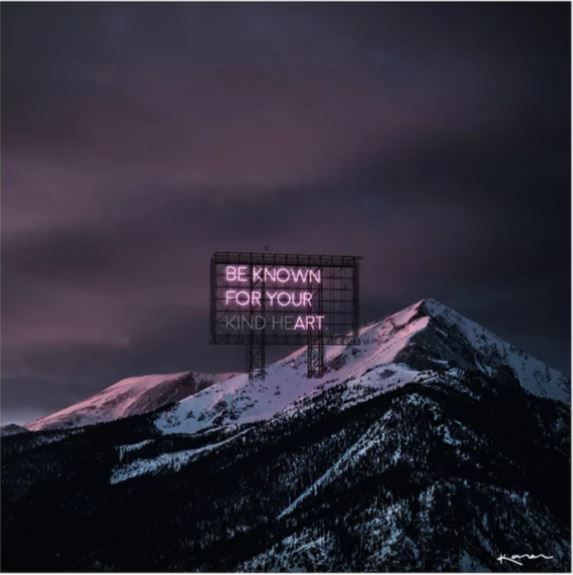 A large billboard with glowing pink neon text reading "BE KNOWN FOR YOUR KIND HEART" stands against a backdrop of snow-capped mountains at dusk, under a dark, cloudy sky. The Kind He(art) by Karen Art Print from Blue Dog Posters captures the mountain peaks dusted with snow and bathed in a soft, pinkish hue.