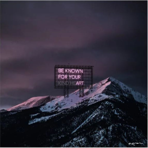 A large billboard with glowing pink neon text reading "BE KNOWN FOR YOUR KIND HEART" stands against a backdrop of snow-capped mountains at dusk, under a dark, cloudy sky. The Kind He(art) by Karen Art Print from Blue Dog Posters captures the mountain peaks dusted with snow and bathed in a soft, pinkish hue.