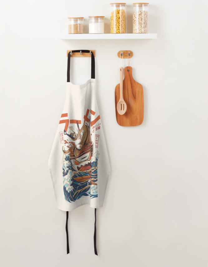 A sublimation-printed white apron with a colorful, artistic design featuring a Japanese-style dragon coiling above ocean waves hangs on a wooden hook. Below a white shelf holding jars of grains, two wooden cutting boards and a wooden utensil are mounted beside it. *The Great Ramen Apron Cream* by *Blue Dog Posters* is machine washable.