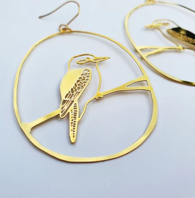 A close-up view of the Denz Kookaburra Dangles Gold Earrings by Blue Dog Posters, showcasing a delicate cut-out design of a Kookaburra perched on a thin branch. Crafted from recycled plated stainless steel, the bird features intricate details on its wings and tail, all framed in elegant oval shapes against a plain white background.