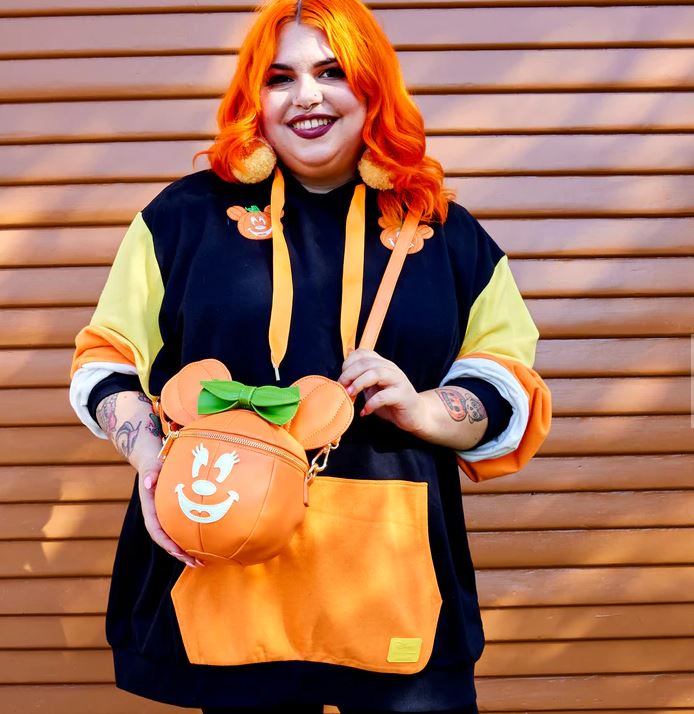 A person with vibrant orange hair is smiling and standing against a wooden backdrop. They are wearing a black and yellow hoodie with orange details and holding a Halloween accessory, specifically the Blue Dog Posters Disney Minnie Pumpkin Face Crossbody Bag. Tattoos are visible on their arms.