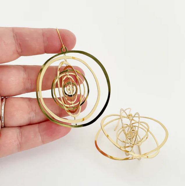 The Denz Orbit Dangles Gold Earrings by Blue Dog Posters are shown, with one gracefully held between fingers and the other resting elegantly on a surface. These lightweight earrings boast a series of concentric, open circular loops crafted from recycled stainless steel, culminating in a dynamic and intricate design.