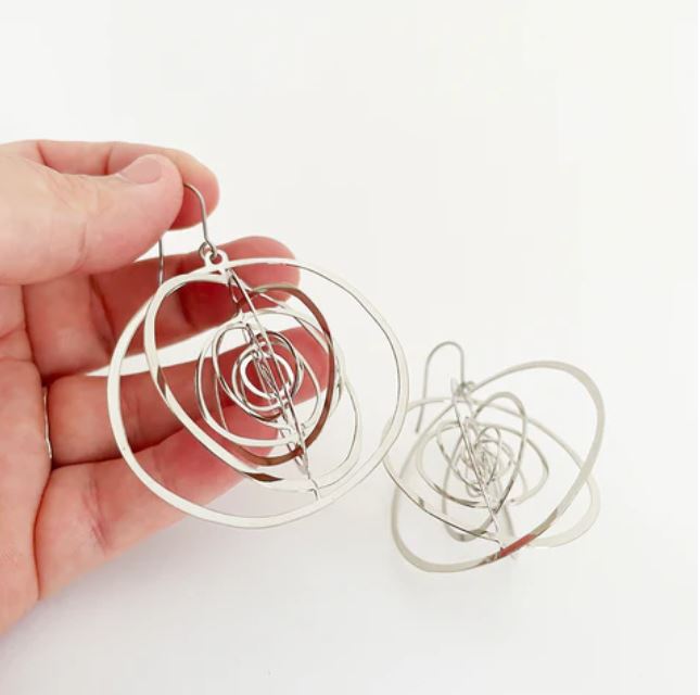 A hand holds a pair of Denz Orbit Dangles Silver Earrings from Blue Dog Posters, made from recycled stainless steel, with the matching earring resting on a white surface. The abstract spiral design is formed from thin, circular metal rings that interconnect to create a geometric pattern. These lightweight earrings bring an elegant charm against the plain white background.