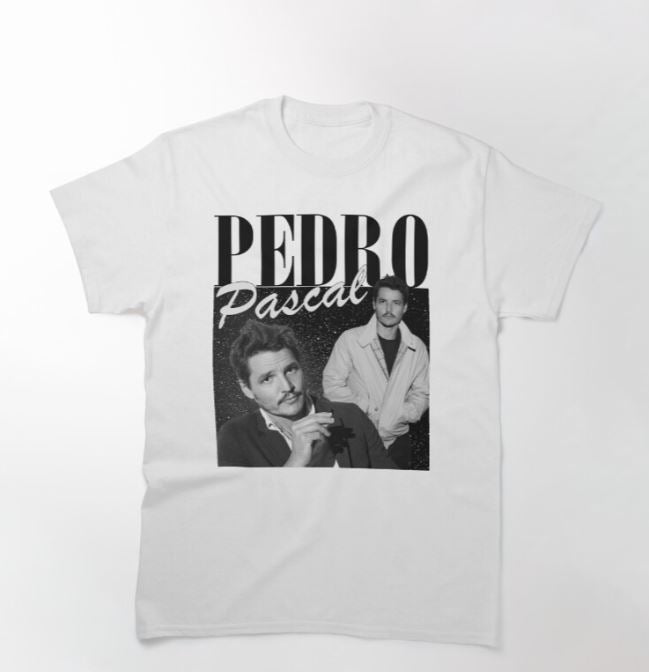 The Pedro Pascal T-shirt White MED from Blue Dog Posters features two black-and-white images of Pedro Pascal, with his name printed in bold, stylized letters above and between the photos. The left image shows him smoking a cigarette, while the right image captures him standing with his hands in his jacket pockets.