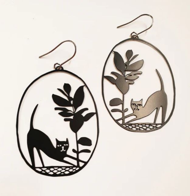 Introducing the Denz Pickles Cat Black Earrings by Blue Dog Posters: a pair of oval-shaped statement earrings with intricate cut-out designs. Each earring features a stretching cat next to a leafy plant, crafted from recycled stainless steel. The black metal emphasizes the delicate and whimsical imagery against a white background.