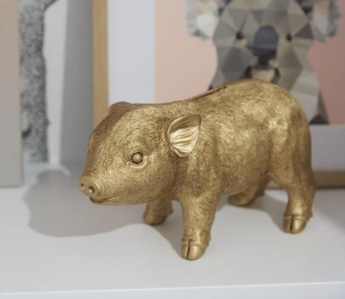 A small, gold-colored Pig Money Box from Blue Dog Posters stands on a white surface. The pig money box has a textured, shiny finish and is placed in front of a framed geometric artwork featuring an animal in the background. The scene exudes a decorative, artistic vibe with the added charm of coin access.