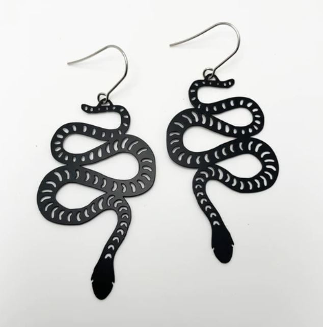 The Denz Snake Black Earrings by Blue Dog Posters are a pair of super lightweight, black, snake-shaped statement earrings with intricate cutout detailing. These earrings depict coiled snakes with distinct heads and tails, attached to simple hook wires. Made from recycled stainless steel, they rest against a plain white background.