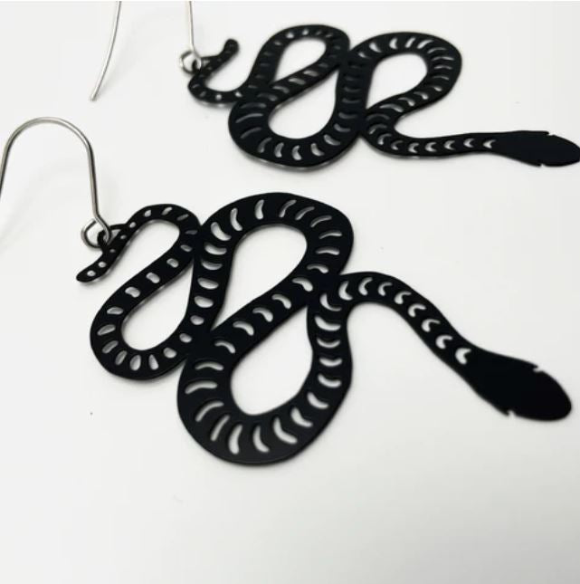 A close-up image of a pair of Denz Snake Black Earrings by Blue Dog Posters, designed to resemble snakes with white patterned markings along their bodies. The super lightweight snakes hang from thin hooks against a plain white background.