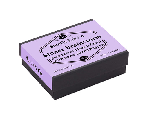 A black-edged rectangular box with a lilac lid features humorous text that reads "Smells Like a Stoner Brainstorm: Pure Genius Ideas Infused With Never Gonna Happen." The packaging, labeled "Hustle and Co Soap - Stoner Brainstorm" by Blue Dog Posters, also highlights "100% Natural Goat's Milk Soap" made in Australia with natural ingredients.