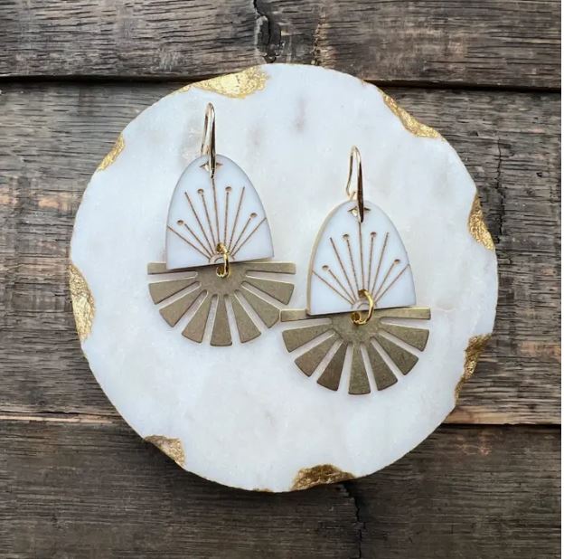 A pair of *Sunrise Sunset White Acrylic & Brass Earrings* by Blue Dog Posters showcased on a round white and gold-trimmed marble plate. The lightweight acrylic earrings feature a white teardrop-shaped piece with golden lines radiating from the center, and below it, a fan-shaped gold metal design with 18K gold plated hooks. The background is wooden.