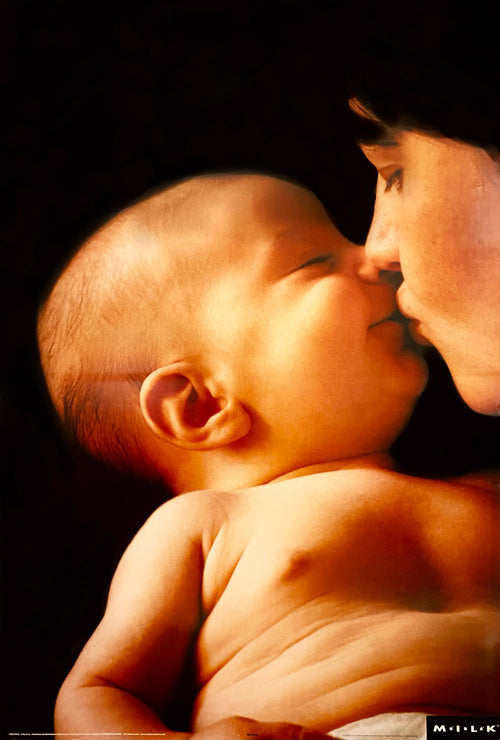 A close-up image called "Baby Kiss" by ALT shows a baby and an adult engaging in a tender moment. The adult, with closed eyes, gently kisses the baby's cheek, evoking a sense of love and care. The dark background highlights their intimate connection. Printed on premium gloss paper, this piece is part of the Scenic Collection with "MILK" written at the bottom right.