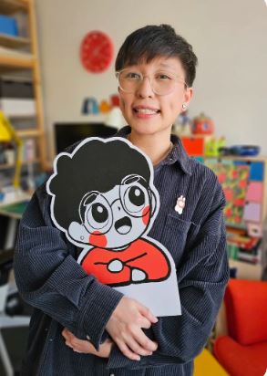 A person wearing glasses and a striped shirt smiles while holding a large cartoon illustration of a character with glasses and curly hair. The background shows a colorful, organized room.