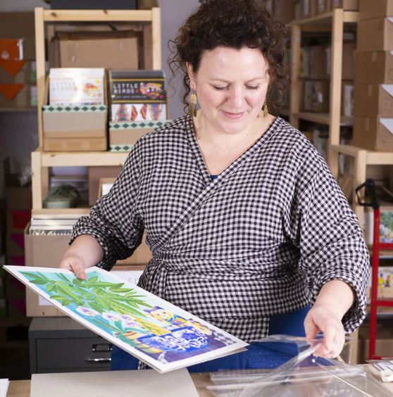 A woman with curly hair, wearing a checked top, smiles while placing a colorful art print into a clear plastic sleeve in a creative studio filled with artwork and supplies.