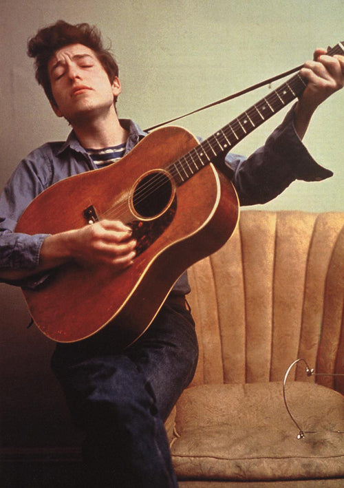 A person with closed eyes, wearing a denim shirt over a striped top, sits on a beige couch strumming the MUS 211 Bob Dylan - Colour Guitar. Their expression appears focused and emotive, while their music collection and a pair of glasses rest beside them on the couch.