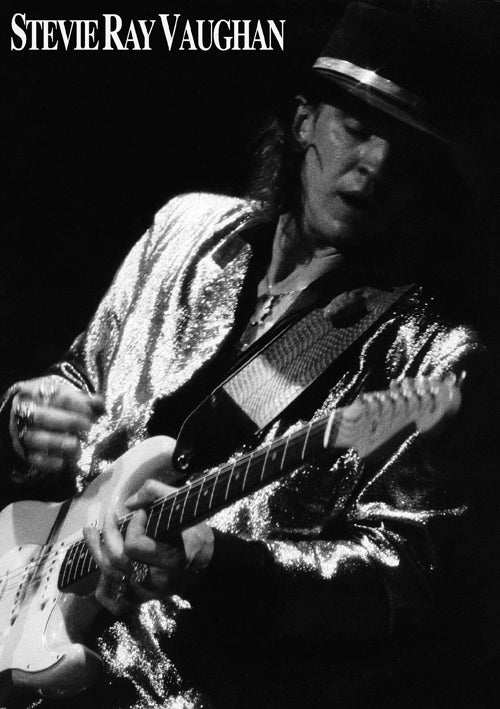 Black and white photo of musician Stevie Ray Vaughn playing an electric guitar. He wears a shiny, metallic jacket and a hat. The image, perfect for any music collection, captures him mid-performance with an intense and focused expression on premium gloss paper from MUS 316's Stevie Ray Vaughn series.