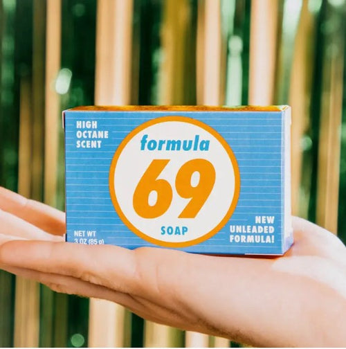 Close-up of hands holding a blue and yellow vintage-style colourful box of "Formula 69 Triple Milled Boxed Bar Soap" by Blue Dog Posters. The box reads "High Octane Scent" and "New Unleaded Formula!" with a net weight of 3 oz (85 g). This Vermont-made, triple-milled soap sits against out-of-focus vertical wooden panels in the background.