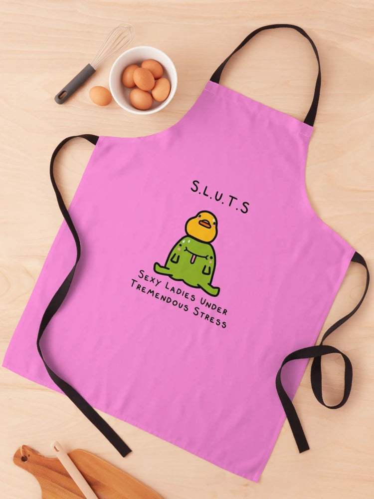 A Blue Dog Posters S.L.U.T.S Apron, featuring a cartoon duck in green clothing and an orange beak with "S.L.U.T.S" above and "Sexy Ladies Under Tremendous Stress" below, is laid flat on a wooden surface. The pink illustrated apron has black straps, with eggs and kitchen utensils from the Homewares Collection positioned nearby.