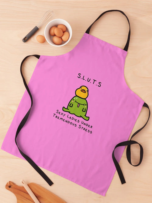 A Blue Dog Posters S.L.U.T.S Apron, featuring a cartoon duck in green clothing and an orange beak with "S.L.U.T.S" above and "Sexy Ladies Under Tremendous Stress" below, is laid flat on a wooden surface. The pink illustrated apron has black straps, with eggs and kitchen utensils from the Homewares Collection positioned nearby.