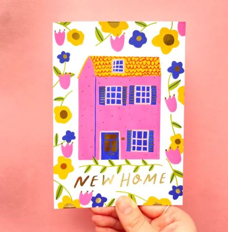 The Blue Dog Posters New Home Pink House Greeting Card features a pink house with blue windows, yellow and purple flowers, "NEW HOME" in gold, comes with a coloured envelope, is FSC certified, and blank inside.