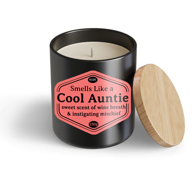 The Cool Auntie Candle Hustle & Co by Blue Dog Posters is a black jar with a wooden lid and a pink label saying, "Smells Like a Cool Auntie: sweet scent of wine breath & instigating mischief." Lid is beside the jar.