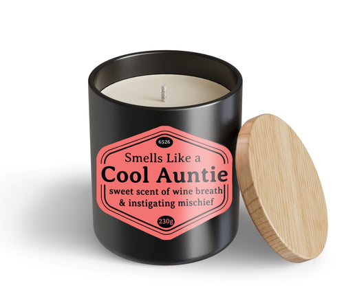 The Cool Auntie Candle Hustle & Co by Blue Dog Posters is a black jar with a wooden lid and a pink label saying, "Smells Like a Cool Auntie: sweet scent of wine breath & instigating mischief." Lid is beside the jar.
