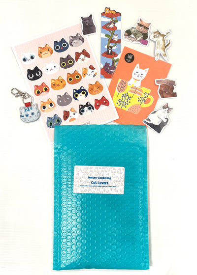 A teal bubble mailer labeled "Mystery Goodie Bag - Cat Lover" by Blue Dog Posters is shown surrounded by cat-themed stickers, postcards, a keychain, and a bookmark with playful cat illustrations on a white background.